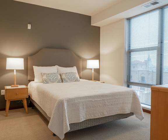 A spacious bedroom with a white, queen-size bed, a dresser, wall art, lamps, decorations, window, and nightstands at Vue Apartment Homes. 