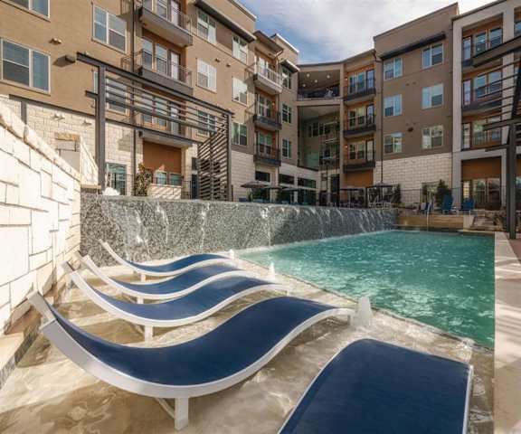 The turquoise outdoor pool with a waterfall cascading into it, wavy navy pool chairs, tables, and chairs with the apartments in the background at Magnolia on the Green Apartments. 