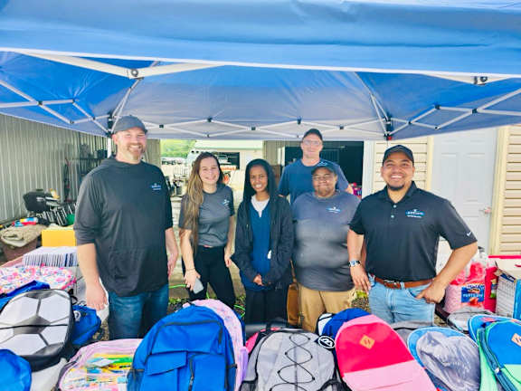 A group of people standing under a blue canopy with various colored backpacks on the ground.