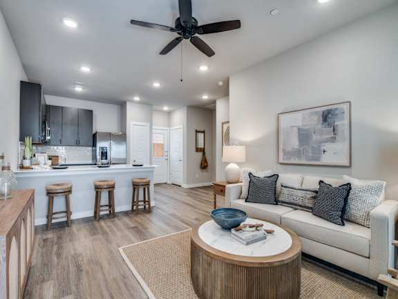 A modern living room with a ceiling fan and a kitchen area in the background.