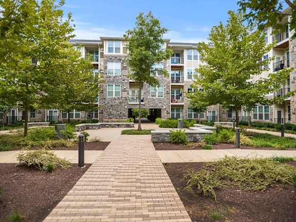 A brick pathway leads to a courtyard with a stone wall and trees in front of apartment buildings.