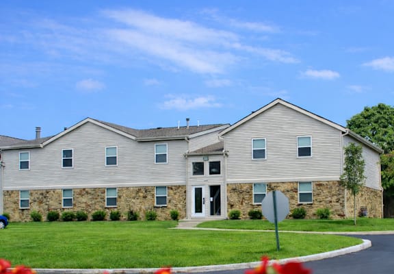 A building with a grey stone facade and a white door is surrounded by a green lawn and red flowers.