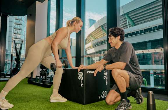 A woman in a beige outfit is doing a squat with a man in a grey t-shirt and black shorts assisting her.