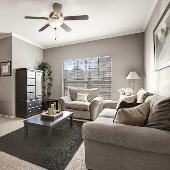 The spacious living room with a light gray couch and plush chair, coffee table, overhead lighting with a ceiling fan, decorations, and view of the dining room with a table, gray chairs, and place settings for four at Stoneleigh on May Apartment Homes. 