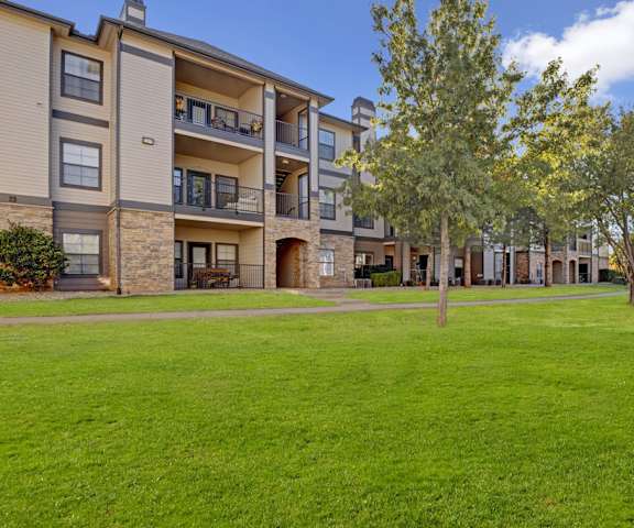 The large, beautiful, grassy area with a pathway and trees behind the tan and stone apartment buildings at Stoneleigh Apartment Homes. 