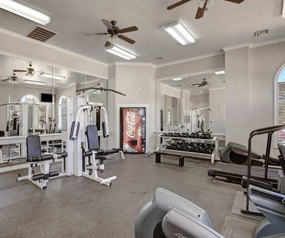  A spacious gym with cardio machines, weight machines, a vending machine, windows, TVs, and weights at Stoneleigh on May Apartment Homes. 