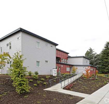A grey building with a red section to the side and a small tree in front.