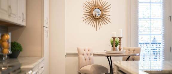 A cozy kitchen nook with a round table, tufted beige chairs, and a decorative sunburst mirror on the wall, complemented by natural light from nearby windows.