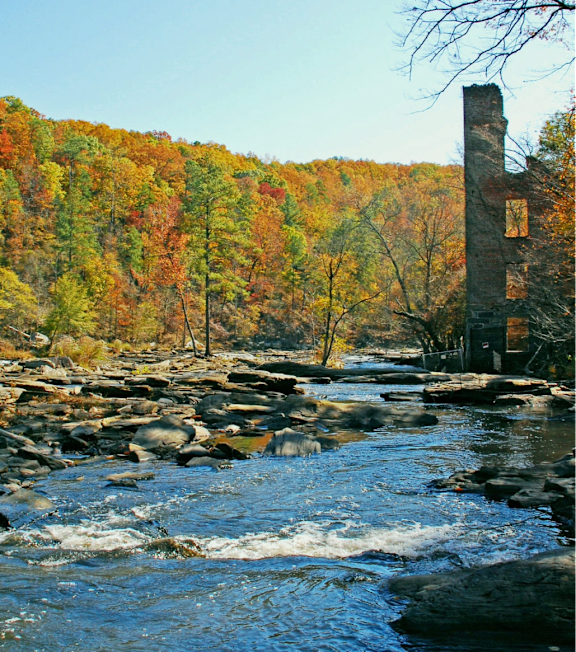A river flows through a forest with a tall, ruined brick chimney in the foreground.