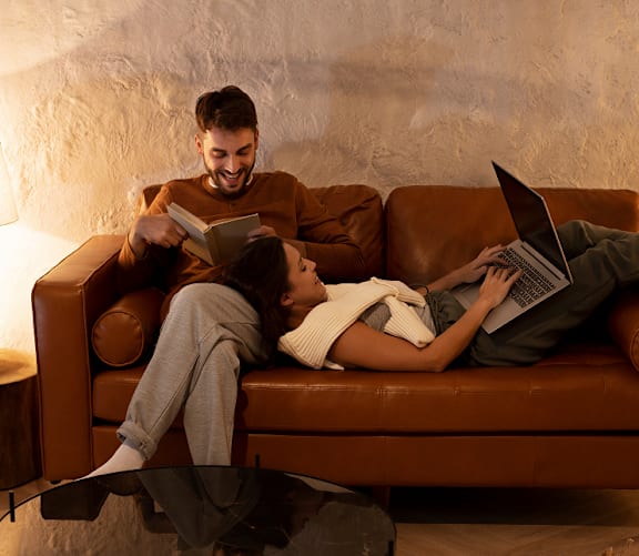 A man and a woman are lounging on a couch with a laptop and a book.