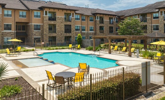 A large swimming pool surrounded by yellow chairs and umbrellas in front of a multi-story apartment building.