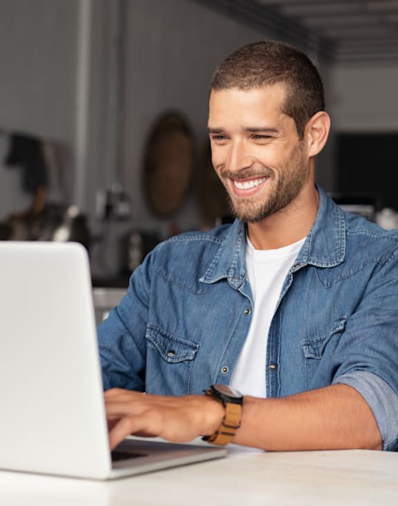 A man in a denim shirt is smiling while using a laptop.