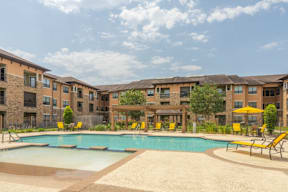 A swimming pool surrounded by yellow chairs and umbrellas in front of apartment buildings.