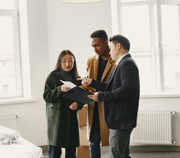 Three people are standing in a room with a bed and a lamp.