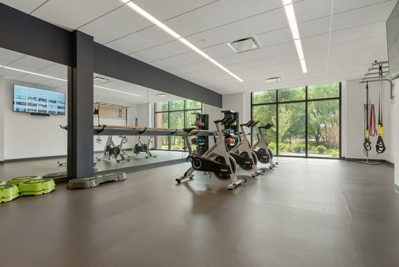 A state-of-the-art fitness center with a row of cardio bicycles, a large mirror, and panoramic windows with views of green space. at Keva Flats, Exton, PA, 19341