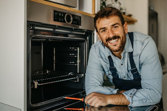 A man with a beard and mustache is wearing a denim shirt and overalls, leaning on an oven in a kitchen.