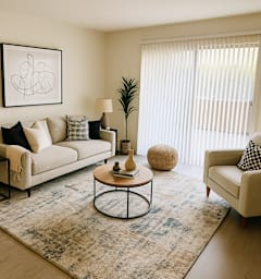 A living room with a beige sofa, a black and white checkered pillow, a round coffee table, a floor lamp, and a framed abstract art piece on the wall.