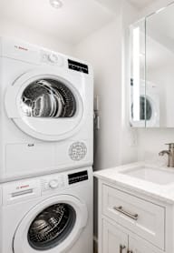 A white washing machine is in a small laundry room with a sink and mirror.