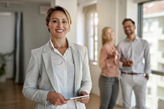 A woman in a white shirt and grey blazer is holding a tablet in a bright room with two other people in the background.
