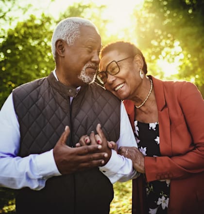 An older couple standing in a park with the man wearing a brown vest and the woman wearing a red blazer.