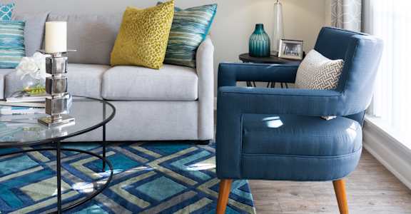 A blue chair sits in a living room with a white couch and a glass coffee table.