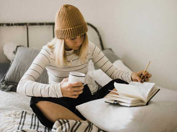a woman sitting on a bed reading a book and drinking coffee