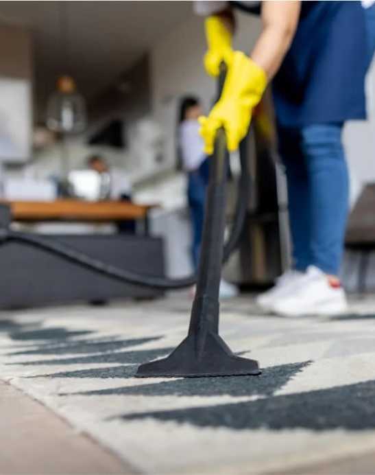 A person in a blue shirt and yellow gloves is vacuuming a rug in a room with other people in the background.