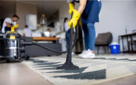 A person in a blue shirt and yellow gloves is vacuuming a rug in a room with other people in the background.