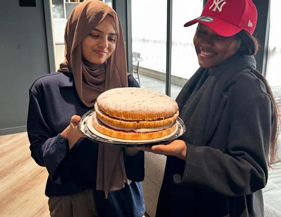 Two women holding a cake in a room with a glass wall.