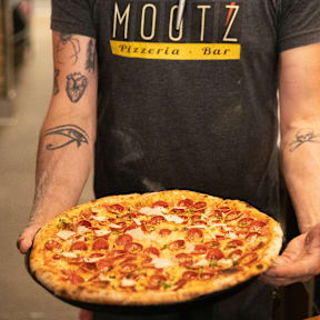 A man in a MOUTZ t-shirt holds a pizza.