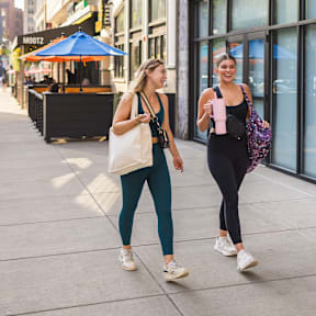 Two women walking on a sidewalk while carrying bags.