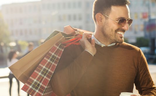 A man in a brown sweater is holding a cup of coffee and shopping bags.