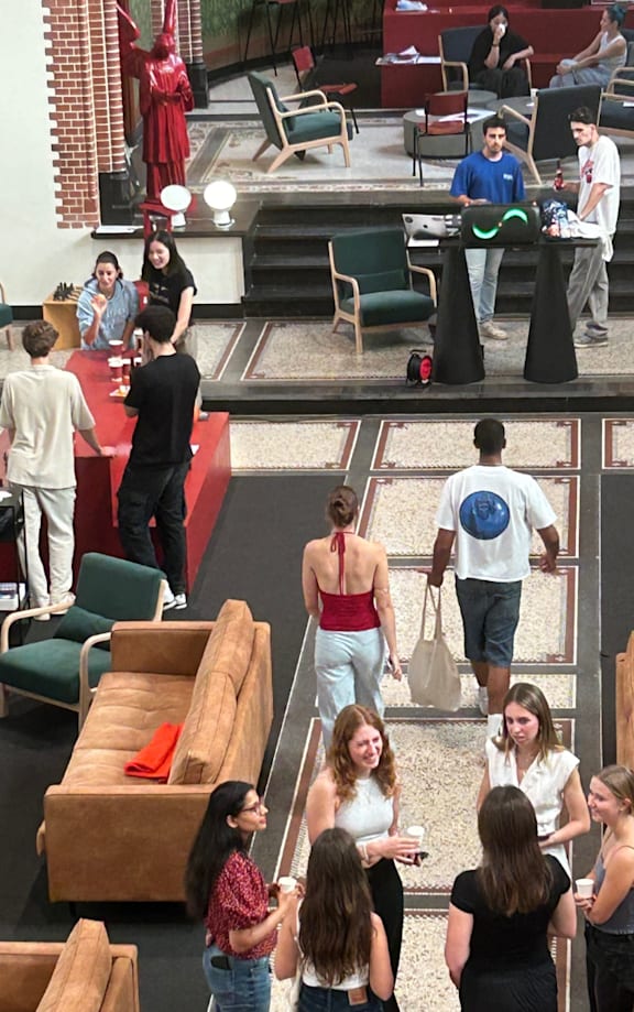 A group of people are gathered in a room with a mix of seating options including sofas, chairs, and a staircase.
