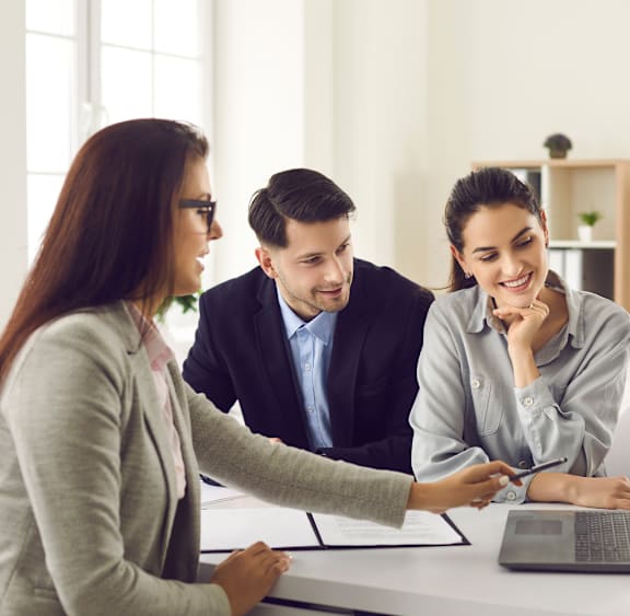 Three people are sitting at a table with papers and a laptop, engaged in a discussion.