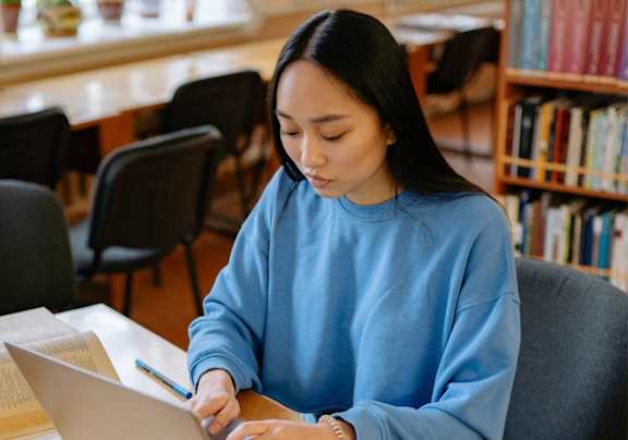 A woman in a blue sweatshirt is working on her laptop at a table with books.