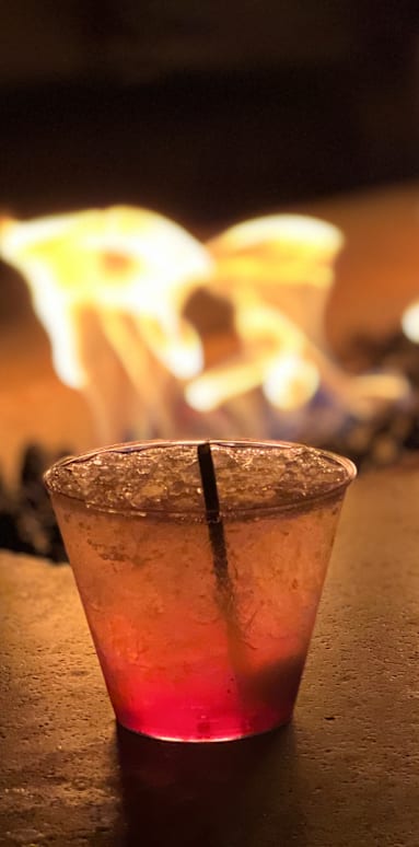 Two glasses of a pinkish drink with a straw in them are in front of a fire.
