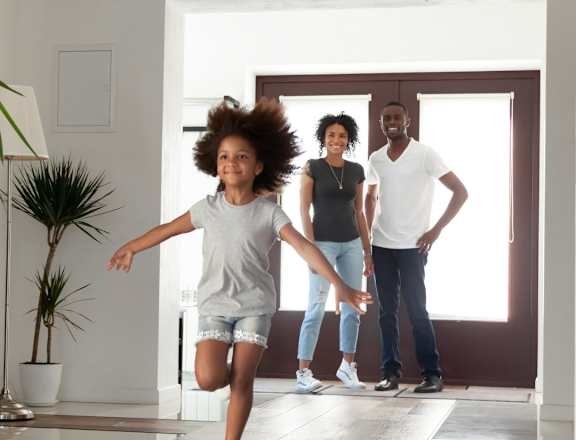 A young girl with an afro is running in a house with a man and a woman standing behind her.