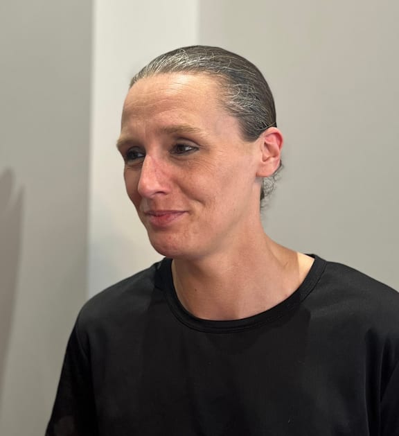 A person with a black shirt is standing against a grey background.