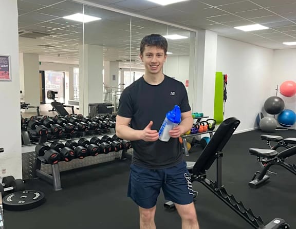 A man in a black shirt and shorts stands in a gym holding a water bottle.