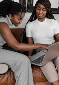Two women are sitting on a couch, one is typing on a laptop at Clarkston Station Apartments, Clarkston