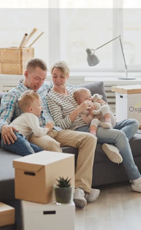 A family of three is sitting on a couch surrounded by moving boxes in a bright room.