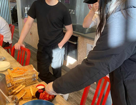 A man in a black shirt stands behind a counter with a woman in a black coat reaching for a bowl of grapes.
