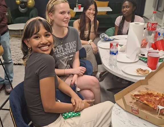 A group of people are sitting around a table with a pizza box in front of them.