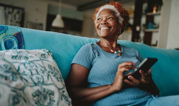 A woman with a joyful expression is sitting on a blue couch, holding a smartphone in her hands.