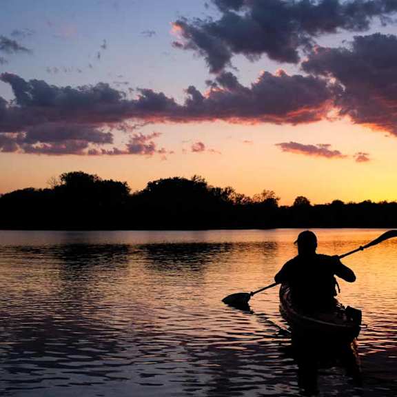 A person is kayaking on a calm body of water during sunset.