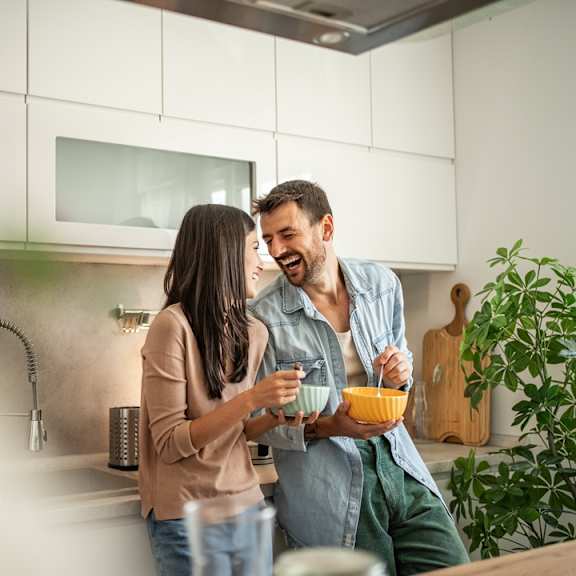 A man and woman are standing in a kitchen, eating cereal together.