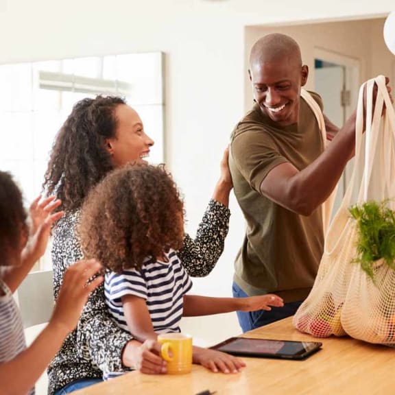 a family of four is gathered around a kitchen table with a grocery bag of vegetables