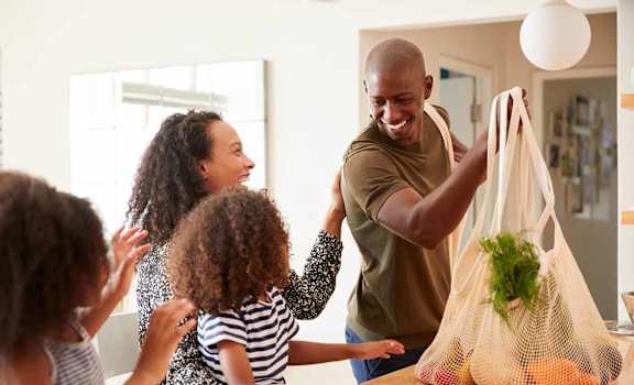 a family of four is gathered around a kitchen table with a grocery bag of vegetables