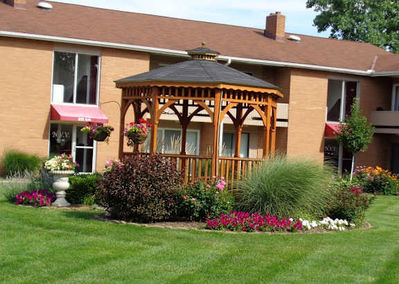 A gazebo is surrounded by a flower garden in front of a brick building.