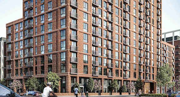 A tall brown building with many windows is surrounded by trees and people walking around.